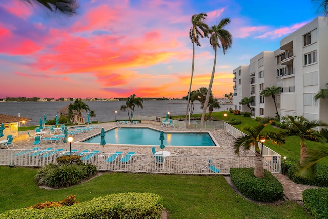 pool at dusk featuring a water view, a patio area, and a lawn