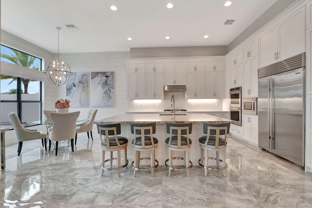 kitchen featuring white cabinetry, built in appliances, light stone countertops, and an island with sink