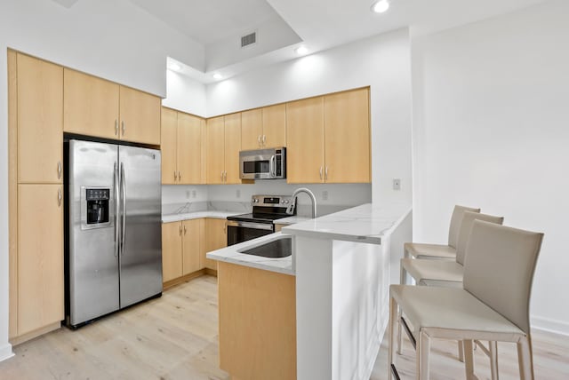 kitchen with light stone countertops, light brown cabinetry, appliances with stainless steel finishes, kitchen peninsula, and light hardwood / wood-style flooring