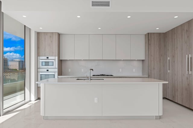 kitchen featuring double oven, a kitchen island with sink, sink, and white cabinets