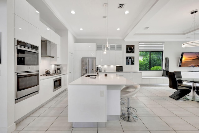 kitchen featuring appliances with stainless steel finishes, wall chimney exhaust hood, decorative light fixtures, white cabinetry, and an island with sink