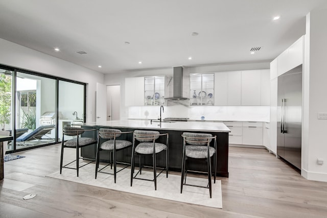 kitchen with wall chimney range hood, white cabinets, a large island with sink, light hardwood / wood-style flooring, and built in fridge