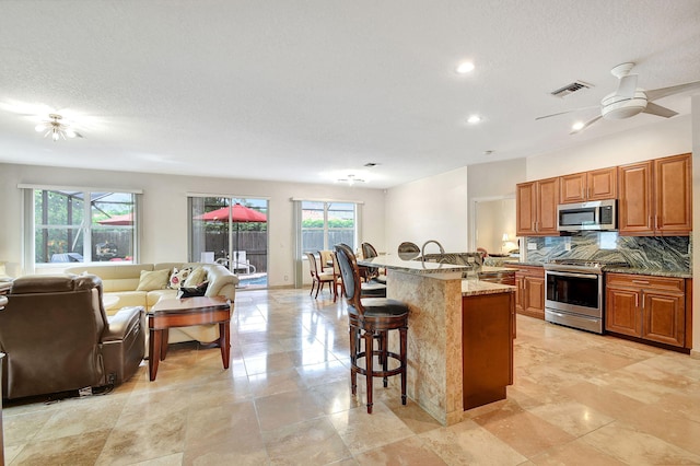 kitchen with a center island with sink, ceiling fan, appliances with stainless steel finishes, a breakfast bar, and light stone counters