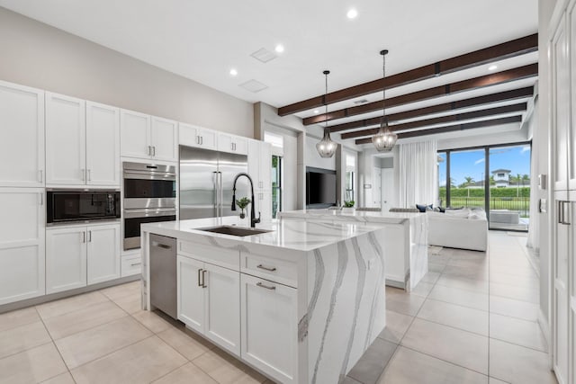 kitchen with light stone countertops, beamed ceiling, decorative light fixtures, built in appliances, and a kitchen island with sink