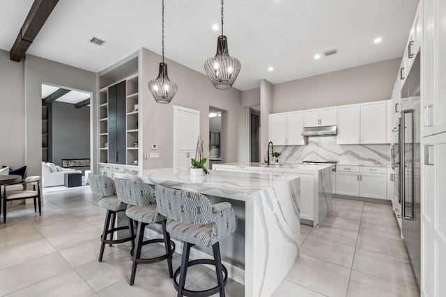 kitchen featuring an island with sink, white cabinetry, light stone countertops, light tile patterned flooring, and pendant lighting