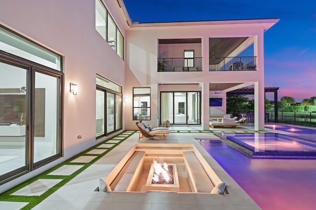 back house at dusk with a patio, a balcony, a swimming pool with hot tub, and a fire pit