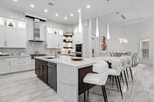 kitchen with wall chimney range hood, white cabinets, sink, and a center island with sink