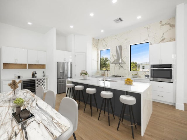 kitchen with wall chimney range hood, beverage cooler, an island with sink, white cabinetry, and stainless steel appliances