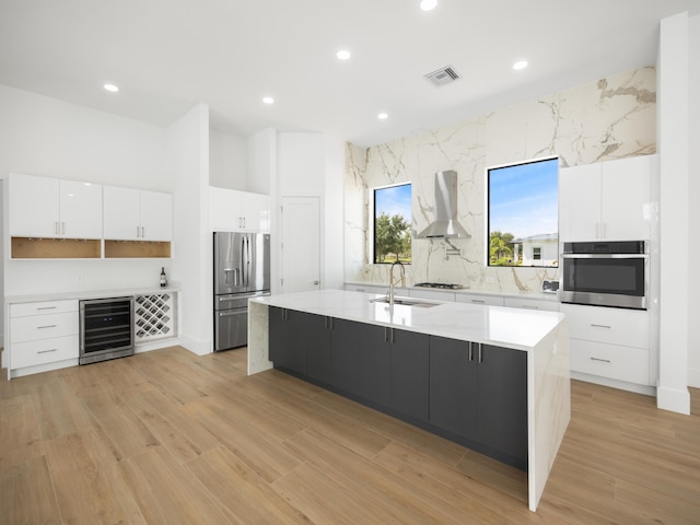 kitchen with a kitchen island with sink, wine cooler, light wood-type flooring, white cabinetry, and appliances with stainless steel finishes