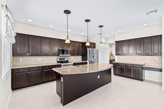 kitchen with dark brown cabinetry, stainless steel appliances, backsplash, pendant lighting, and a center island with sink