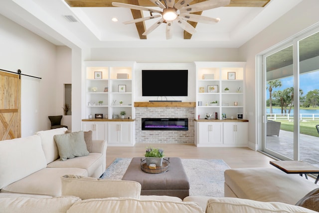 living room featuring a brick fireplace, a raised ceiling, ceiling fan, a barn door, and light hardwood / wood-style flooring