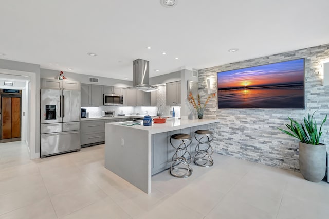 kitchen with light tile patterned floors, appliances with stainless steel finishes, a kitchen bar, island range hood, and gray cabinetry