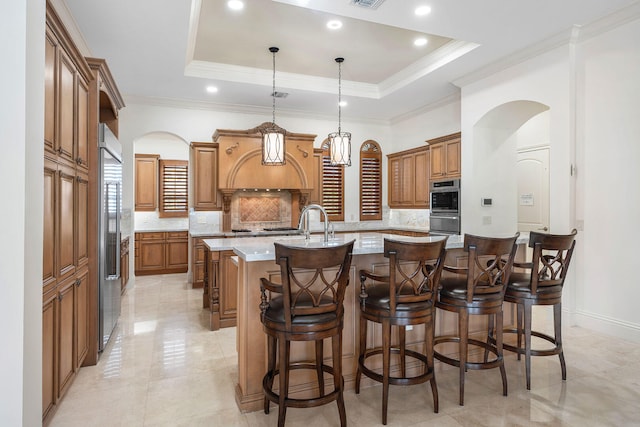 kitchen with a raised ceiling, a center island with sink, light stone counters, and hanging light fixtures