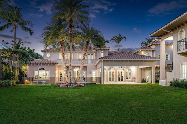 back house at dusk with a lawn, a balcony, a swimming pool, and french doors