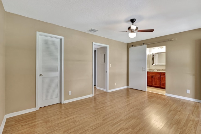 unfurnished bedroom featuring connected bathroom, light hardwood / wood-style flooring, a barn door, a textured ceiling, and ceiling fan