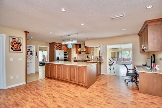 kitchen with light stone countertops, light wood-type flooring, a kitchen island, stainless steel appliances, and wall chimney exhaust hood