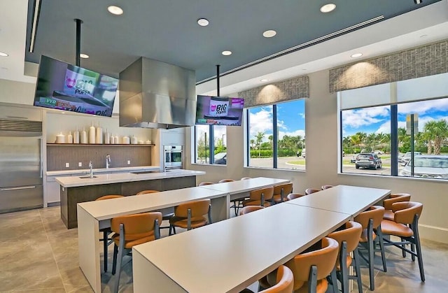 kitchen featuring wall chimney exhaust hood, light tile patterned floors, an island with sink, appliances with stainless steel finishes, and a breakfast bar area