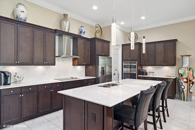 kitchen with stainless steel appliances, sink, wall chimney exhaust hood, decorative light fixtures, and a kitchen island with sink