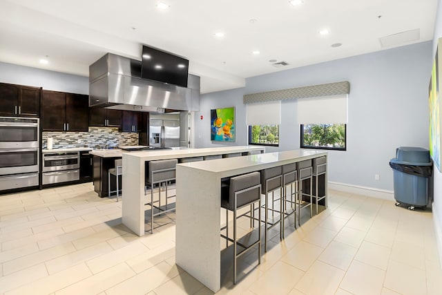 kitchen with a kitchen bar, stainless steel appliances, decorative backsplash, an island with sink, and wall chimney range hood