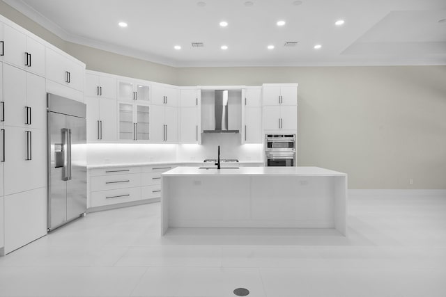 kitchen with wall chimney range hood, a center island with sink, white cabinetry, appliances with stainless steel finishes, and ornamental molding