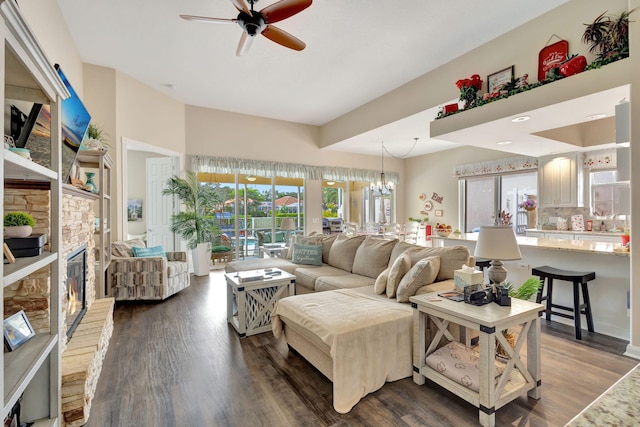 living area featuring ceiling fan, dark wood-style flooring, a stone fireplace, and a chandelier