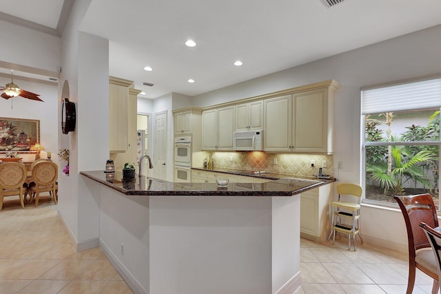 kitchen with kitchen peninsula, cream cabinetry, white appliances, and dark stone counters