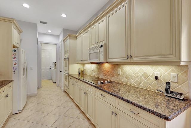 kitchen with white appliances, light tile patterned flooring, cream cabinetry, dark stone counters, and decorative backsplash