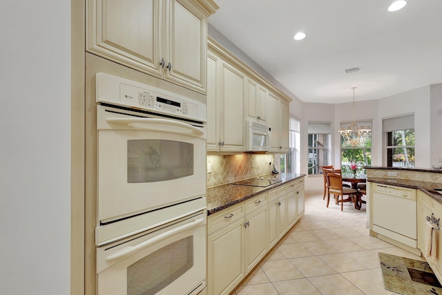 kitchen featuring white appliances, cream cabinetry, dark stone counters, pendant lighting, and an inviting chandelier