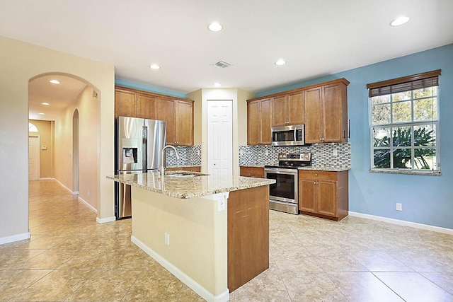 kitchen featuring a center island with sink, appliances with stainless steel finishes, tasteful backsplash, light stone counters, and sink