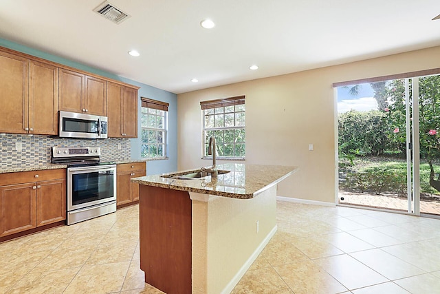 kitchen featuring a center island with sink, stainless steel appliances, decorative backsplash, light stone counters, and sink