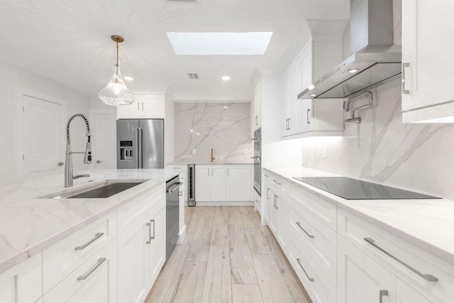 kitchen featuring a skylight, stainless steel appliances, sink, wall chimney range hood, and white cabinetry