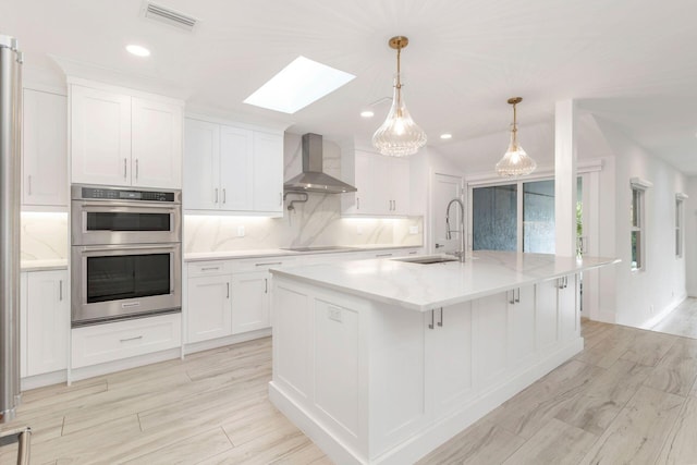 kitchen with sink, an island with sink, white cabinetry, and wall chimney range hood