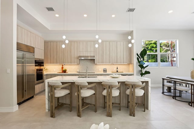 kitchen with light stone countertops, light brown cabinets, decorative light fixtures, a center island with sink, and appliances with stainless steel finishes