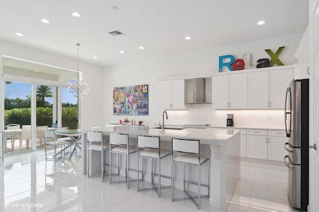 kitchen featuring stainless steel refrigerator, wall chimney exhaust hood, a center island with sink, and white cabinets