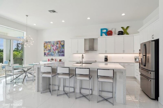 kitchen with wall chimney range hood, sink, stainless steel fridge, white cabinetry, and a center island with sink