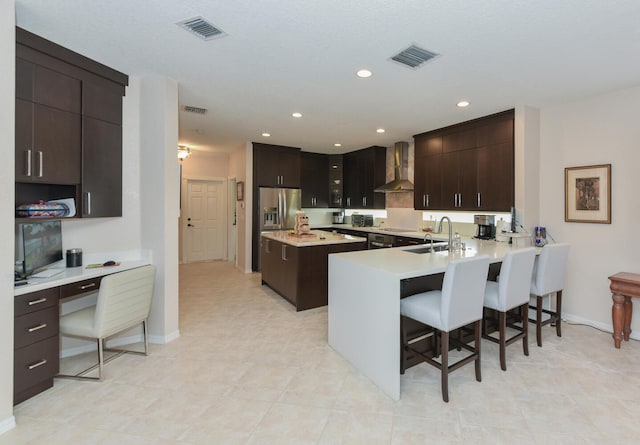 kitchen with kitchen peninsula, wall chimney exhaust hood, a kitchen bar, and stainless steel refrigerator with ice dispenser