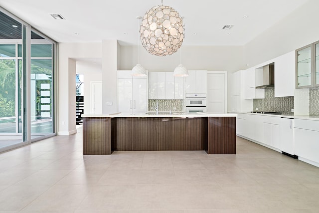 kitchen with white cabinetry, wall chimney exhaust hood, an island with sink, white appliances, and hanging light fixtures