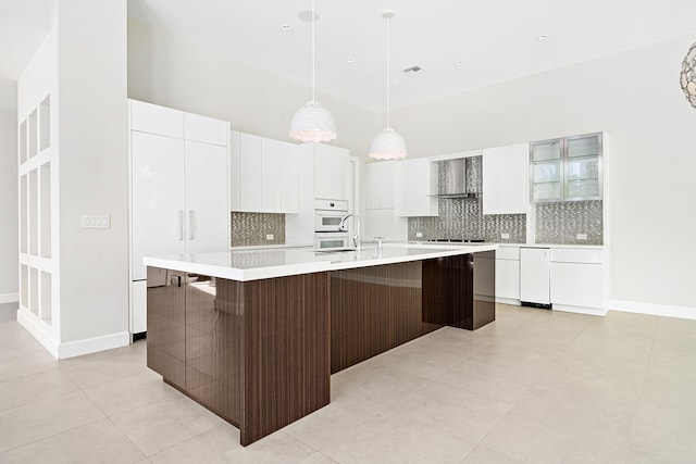 kitchen featuring tasteful backsplash, dark brown cabinetry, an island with sink, white cabinets, and wall chimney exhaust hood