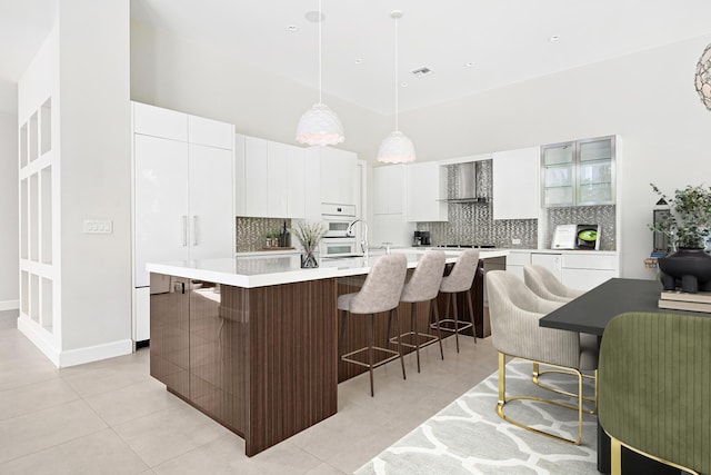 kitchen featuring white cabinets, a breakfast bar area, a kitchen island with sink, wall chimney range hood, and pendant lighting