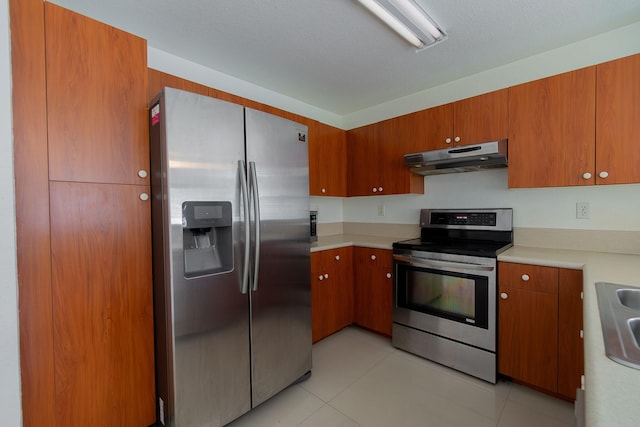 kitchen with appliances with stainless steel finishes, brown cabinets, light countertops, light tile patterned floors, and under cabinet range hood