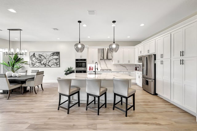 kitchen with stainless steel appliances, wall chimney exhaust hood, decorative light fixtures, white cabinets, and a center island with sink