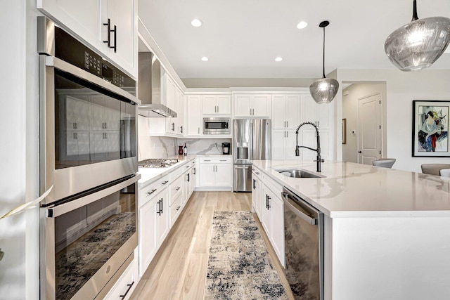 kitchen featuring hanging light fixtures, an island with sink, appliances with stainless steel finishes, wall chimney exhaust hood, and sink