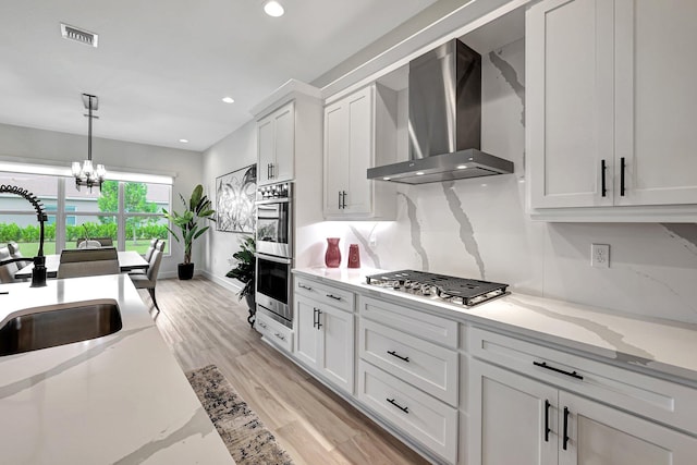 kitchen featuring wall chimney exhaust hood, light hardwood / wood-style flooring, stainless steel appliances, a notable chandelier, and white cabinets