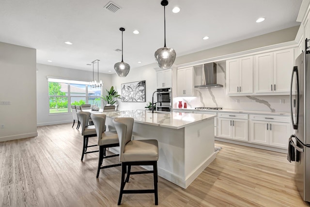 kitchen with a kitchen island with sink, wall chimney range hood, hanging light fixtures, stainless steel appliances, and white cabinetry