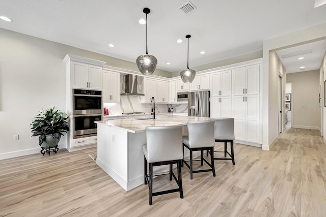 kitchen featuring a kitchen island with sink, wall chimney range hood, stainless steel appliances, pendant lighting, and white cabinetry