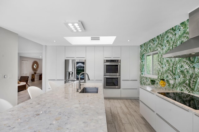 kitchen with wall chimney range hood, sink, white cabinets, appliances with stainless steel finishes, and light hardwood / wood-style floors