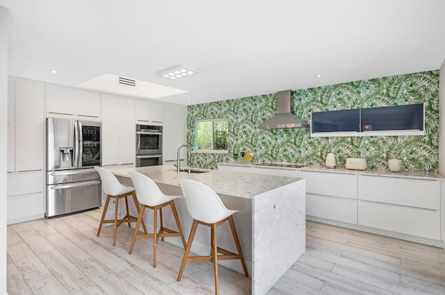 kitchen with sink, white cabinetry, stainless steel appliances, wall chimney exhaust hood, and a kitchen island with sink