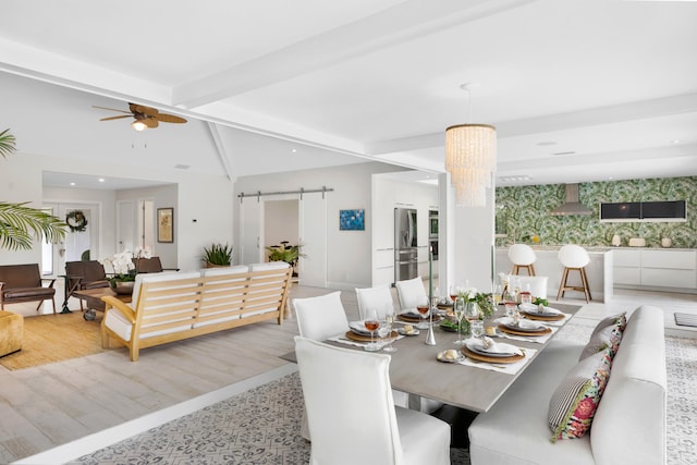 dining area with ceiling fan with notable chandelier, beamed ceiling, light wood-type flooring, and a barn door