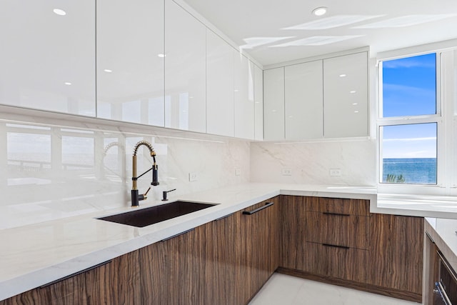 kitchen featuring sink, backsplash, white cabinets, a water view, and light stone counters