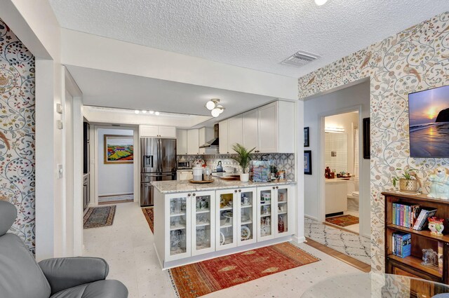 kitchen with white cabinetry, wall chimney range hood, light stone counters, stainless steel refrigerator with ice dispenser, and a textured ceiling